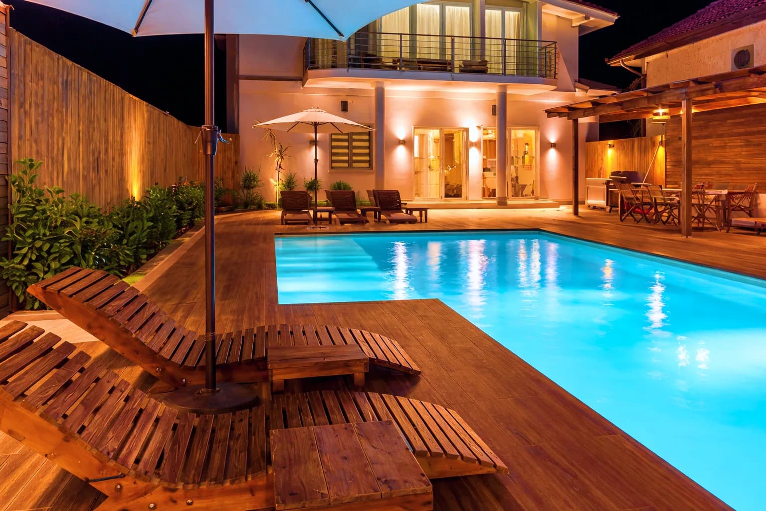 Salon extérieur en bois au bord d’une piscine, de nuit